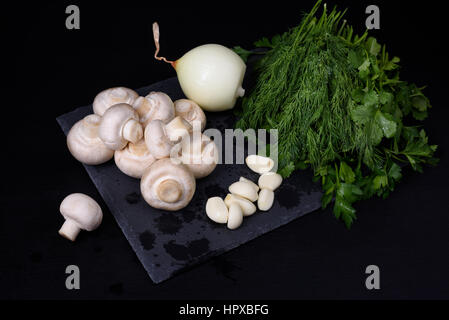 Junge Frühling Gemüse auf Schiefer, schwarzer Hintergrund. Champignon, Pilze, Knoblauch, Zwiebel, Dill, Petersilie und Koriander gewaschen und vorbereitet für Koch Stockfoto
