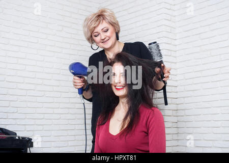 Schöne Frau an der Friseur Föhnen ihre Haare. Trocknen die Haare mit Trockner und runden Haarbürste.  Glückliche junge Frau und ein Friseur mit Ventilator maki Stockfoto