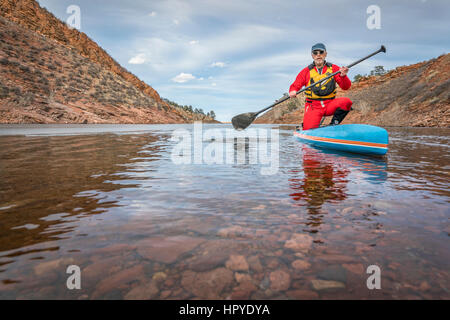 Senior männlichen Paddler in Trockentauchanzug ist Stand-up Paddeln auf See in Colorado, Winterlandschaft mit etwas Eis genießen. Stockfoto