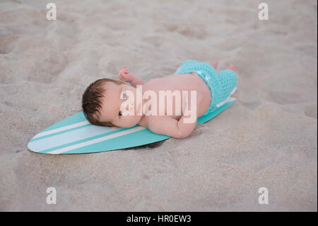 Zwei Wochen altes neugeborenes Baby junge auf einem winzigen, türkis blau-weißen Surfbrett liegend. Er trägt Aqua gefärbt, Boardshorts und liegt an einem Sandstrand