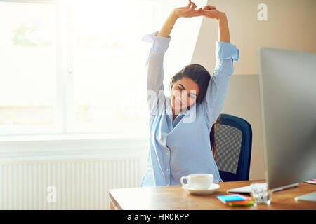 Grinsende junge Frau in blau Langarm-Shirt dehnen vor Schreibtisch zu Hause Stockfoto