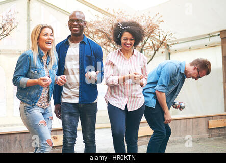 Gruppe von vier Jugendlichen Petanque Spieler tragen legere Kleidung stehend halten Metall Kugeln in die Hände spielen und lachen, Stockfoto