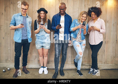 Gruppe von Jugendlichen auf Party oder Tagung tragen legere Kleidung stand in der Nähe von Holzwand alle starrte auf ihre Smartphones, alle Lächeln auf den Lippen Stockfoto