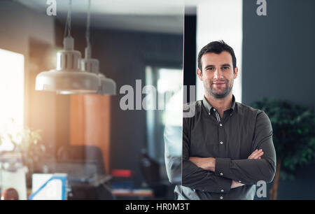 Porträt eines selbstbewusst und erfolgreich jungen Geschäftsmann lächelnd stehend mit seinen Armen überquerte in einem modernen Büro Stockfoto