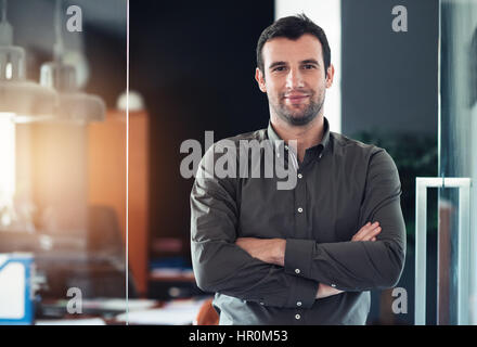 Porträt eines selbstbewusst und erfolgreich jungen Geschäftsmann lächelnd stehend mit seinen Armen überquerte in einem modernen Büro Stockfoto