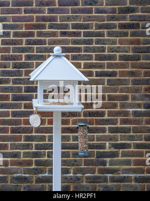 Vogelhaus, neu gesetzt vor einer Wand und mit einem Mutterhalter und Kokos und Samen auf dem Tisch.  Das Vogelhaus hat eine weiße Spitzdach. Stockfoto