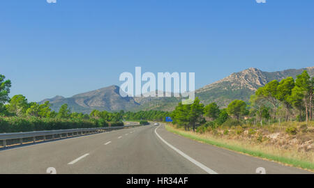 Sonnenschein am Coastal Highway.  Einsames weißes Auto fährt entlang durch Ausläufer und Gebirgsketten an den Rändern des europäischen Kontinents im ländlichen Spanien. Stockfoto