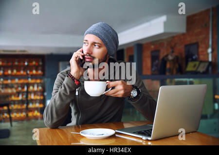 Junge hübsche Hipster Mann mit Bart sitzen im Café, Handy, im Gespräch mit Tasse Kaffee. Laptop auf Holztisch. Stockfoto