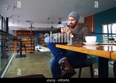 Junge hübsche Hipster Mann mit Bart SMS mit seinem Mobiltelefon an der Bar und haben einen Cappuccino. Stockfoto