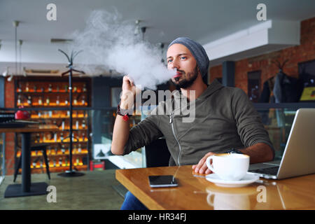 Junge hübsche Hipster Mann mit Bart sitzen im Café bei einer Tasse Kaffee, dampfen und Veröffentlichungen eine Wolke aus Dampf. Arbeiten am Laptop und eine kleine Pause. Mit Textfreiraum. Stockfoto