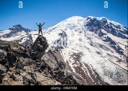 Ein Mann der erhobenen Arme Victory-Zeichen oben auf einem Felsvorsprung mit Mt Rainier und Gletschern im Hintergrund über ihm droht. Anfang der 3. Burrough Stockfoto
