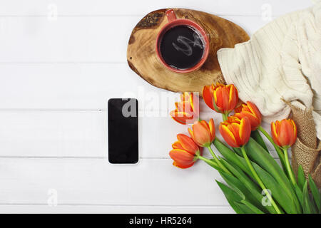 Overhead Schuss eine Telefon mit einer heißen Tasse Kaffee und ein Blumenstrauß mit einer gemütlichen stricken Tulpe Decke über weiße Holz Tischplatte werfen. Flach legen Top v Stockfoto