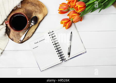 Overhead Schuss eine Tasse Kaffee, Ernährungstagebuch, stricken Decke und einen Blumenstrauß orange und gelbe Tulpe über weiße Holz Tischplatte werfen. Flach legen t Stockfoto