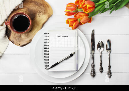 Overhead Schuss ein Ernährungstagebuch über leere Teller mit einer Tasse Kaffee und ein Blumenstrauss Frühling Tulpe über weiße Holz Tischplatte. Flach legen t Stockfoto