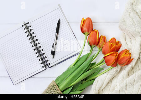 Overhead schoss ein Bouquet von orange und gelbe Tulpen und eine gemütliche Decke über weiße Holz Tischplatte mit ein offenes Buch und Stift bereit, Zeitschrift stricken Stockfoto