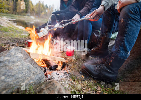 Untere Partie der jungen Freunde rösten Marshmallows über dem Lagerfeuer am Seeufer Stockfoto