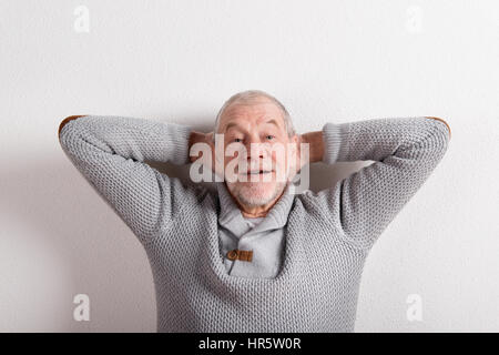 Gut aussehend senior lächelnder Mann in grauen Pullover, die Hände hinter dem Kopf. Studio gegen weiße Wand erschossen. Stockfoto