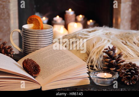 Lesung durch eine Kerze beleuchtet offenen Kamin in einem gemütlichen englischen Haus mit einer Tasse heißen Tee und eine Decke (werfen) im Herbst/Frühwinter Stockfoto