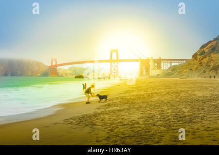 Mann spielt mit seinem Hund in Baker Beach bei Sonnenuntergang mit Golden Gate Bridge. Touristen zu Fuß am Ufer. Urlaub, Reisen und Freizeit in San Francisc Stockfoto