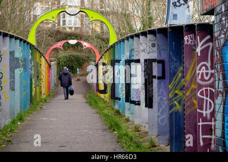 Ein Mann geht über eine Fußgängerbrücke auf einer Eisenbahnlinie in Montpelier, Bristol, die geschmückt mit bunten Streetart auf seine Rechtschreibung aus tiefen Metall-Lamellen Stockfoto