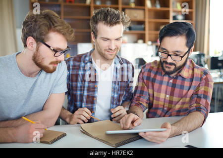 Team von jungen Unternehmern bei der Arbeit: drei junge ehrgeizige Männer sitzen am Tisch Planungsstrategie, neue Geschäftsidee, asiatischen Mann zeigte auf Tablet Stockfoto