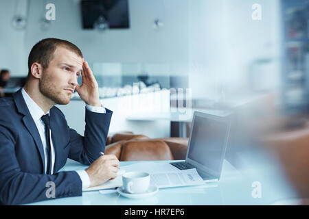 Hübscher junger Geschäftsmann tragen formelle Kleidung arbeiten während der Kaffeepause im modernen Speisesaal: Suchen von Dokumenten mit müde und du Stockfoto
