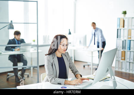 Mehrere Geschäftsleute beschäftigt im Fokus arbeiten in modernen Büro am Schreibtisch mit Computer, junge asiatische Frau, die vorn etwas eingeben Stockfoto