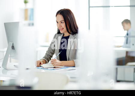 Porträt der erfolgreiche junge Geschäftsfrau beschäftigt, arbeiten am Schreibtisch im modernen Büro, Notizen im Planer und Computer-Bildschirm betrachten Stockfoto