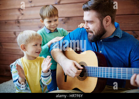 Bärtige Kindergärtnerin Musikstunde zu kleinen Jungen geben: er spielt Gitarre und singt ein lustiges Lied während seiner Schüler mit Interesse auf ihn zu hören Stockfoto