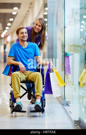 Behinderte jungen Mann im Rollstuhl, die gute Zeit mit seinen lieben Freundin in der Mall, Wandern mit Einkaufstüten und fröhlich lächelnd Stockfoto