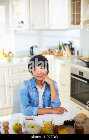Schöne junge Frau in Jeanshemd Blick in die Kamera Lächeln beim Frühstück in modernen Beige Küche Stockfoto