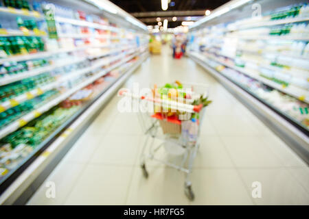 Verschwommene Einkaufswagen voller Flaschen mit Wasser, Obst und Gemüse stehen zwischen zwei Kühlschränke mit Milchprodukten Stockfoto