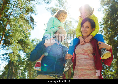 Lächelnde Paar mittleren Alters, Blick in die Kamera während des Tragens ihre niedlichen kleinen Töchter auf Schultern, lens flare Stockfoto