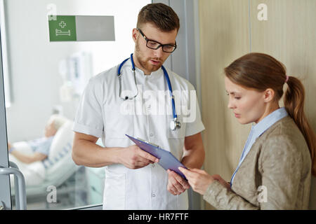 Junger Arzt zeigt Patienten medizinischen Bericht, junge Frau wartet außerhalb geschlossener Ward, erklären den Krankheitsfall von ihrem älteren relative d verärgert Stockfoto