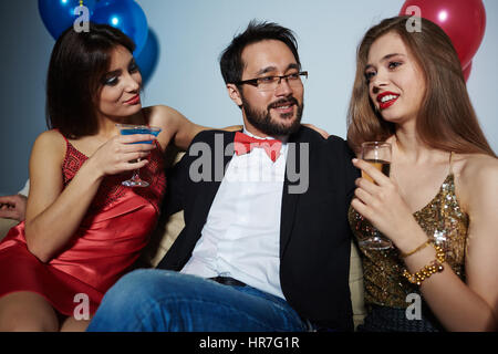 Bärtige asiatischen Geschäftsmann mit Fliege etwas umarmen zwei hübsche Frauen beim sitzen zwischen ihnen, sie haben gute Zeit in Nachtclub Stockfoto