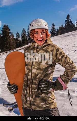 Oberkörper-Aufnahme bei sonnigem Wetter eine rothaarige, lachende Frau mit Helm, stehend auf der Piste mit Snowboard in seiner Hand. Stockfoto