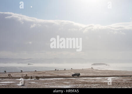 Malerische Aussicht auf den Baikalsee im Winter während eines Schneesturms und ein schwarzes Auto fährt entlang der felsigen Küste. Stockfoto