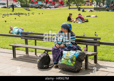 Eine östliche europäische Frau ein Kopftuch sitzen auf einer Bank in einem Park in London mit ihrem Gepäck. Stockfoto