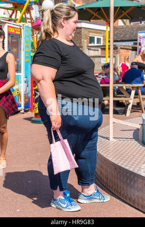 Eine übergewichtige Frau, die neben einer Fahrt auf einem Rummelplatz hält ein kleines Kind Rosa Tasche stand. Stockfoto