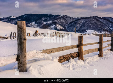 schöne warme Winter Sonnenaufgang am bewölkten Morgen Schnee bedeckten Felder mit hölzernen Zaun in der Nähe von ländlichen Gebiet der Karpaten Stockfoto