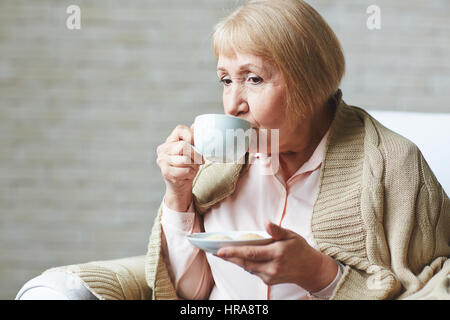 Nachdenklich Seniorin sitzt auf Sessel mit Tasse Kaffee und bedeckte Schultern mit warmen Plaid, Taille-Up portrait Stockfoto
