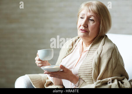 Melancholische senior Frau mit Karomuster auf Schultern halten Kaffeetasse in einer Hand und Untertasse mit Cookies in anderen beim Entspannen im Wohnzimmer Stockfoto