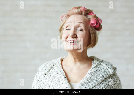 Pensionierte Frau mit Rollen in Haaren und geschlossenen Augen stehen auf weißen Hintergrund und Zukunft träumen Stockfoto