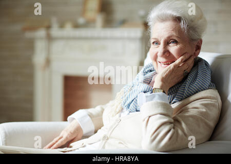 Lächelnd Seniorin mit natürlichen Make-up wegsehen sitzend auf gemütlichen Sessel, Kamin Stockfoto