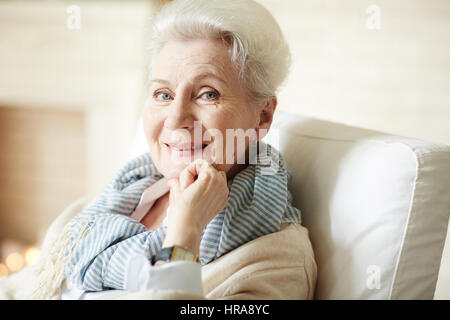 Porträt der schöne alte Dame in Beige Strickjacke und gestreiften Schal im Sessel sitzen, Blick in die Kamera und fröhlich lächelnd Stockfoto
