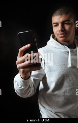 Hübscher afroamerikanische junger Mann Athlet in Hoodie mit Handy auf schwarzem Hintergrund Stockfoto