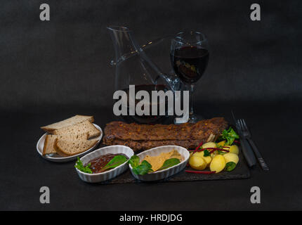 Rack mit Schweinerippchen mit Kartoffeln, Soßen, Brot und Wein in eine Karaffe und Glas, exquisiten dinner Stockfoto