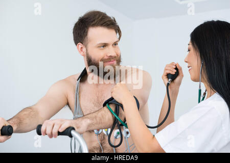Ärztin, die Messung des Blutdrucks von männlichen Patienten Radfahren mit Elektrostimulator Elektroden am Körper im Krankenhaus Stockfoto