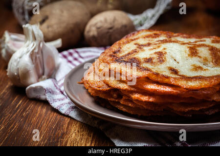 Stapel von Kartoffel Pfannkuchen auf einem Holztisch. Im Hintergrund Kartoffeln, Zwiebeln und Knoblauch Stockfoto