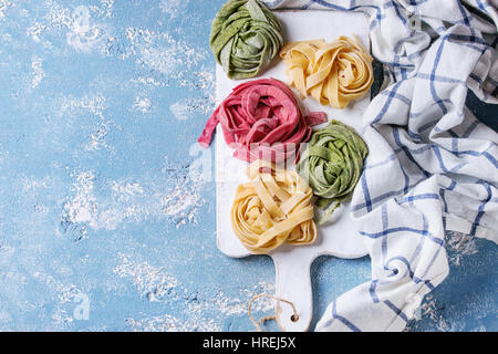 Verschiedene farbige frische rohe hausgemachte Teigwaren Tagliatelle grün Spinat, rosa rote Beete und gelb auf weiß Holzbrett über blaue concr Stockfoto